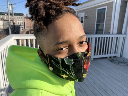 A black woman with locs wearing a neon green hoodie with a black mask with embroidered red and pink flowers and green leaves on it.