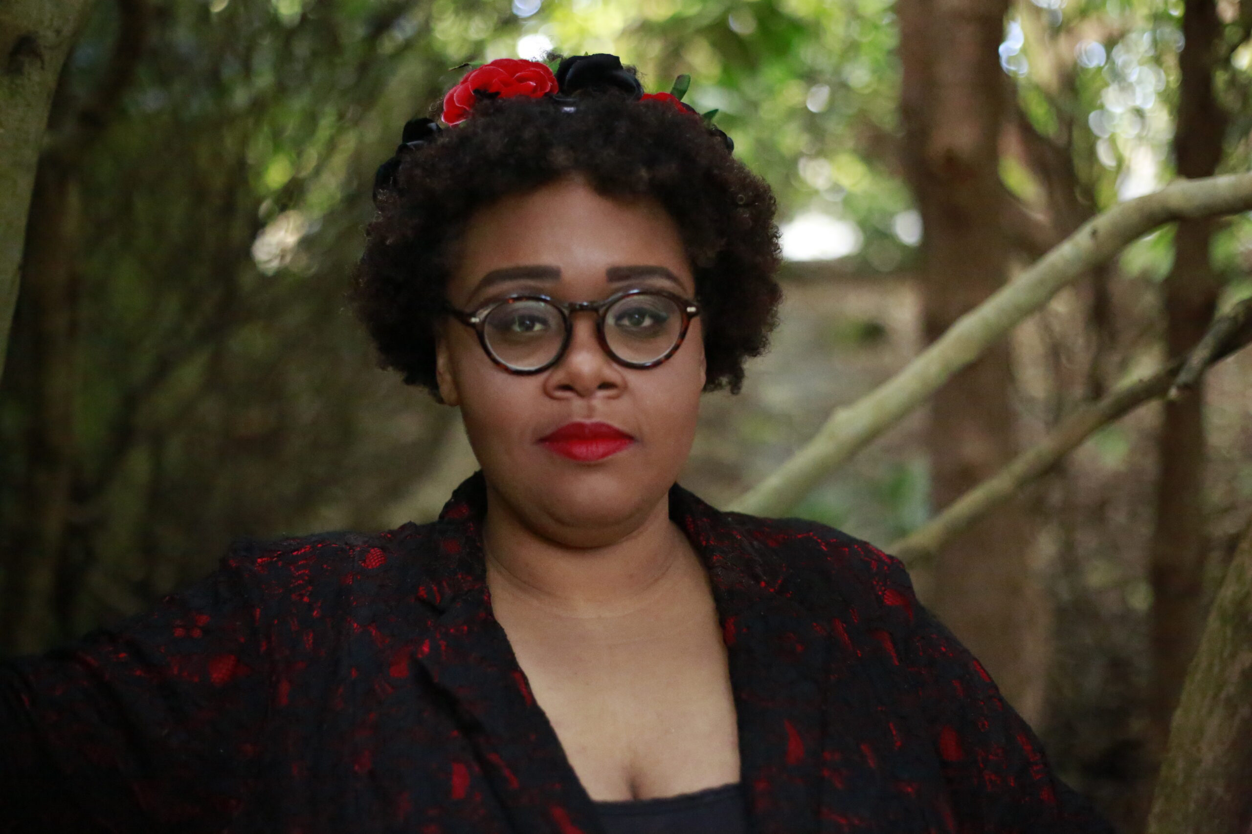 A portrait of Darienne standing in front of some tree. She is wearing a red and black floral crown and a black and red lacy blazer. Her fully made up face is unsmiling but features kind eyes