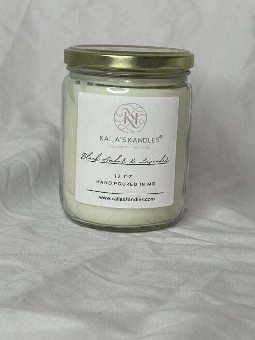 a clear glass jar with a gold top. Inside is filled with fragrant white candle wax. The front has a logo for Kaila's Kandles with the scent black amber and lavender listed on the front as well as 12 ounces