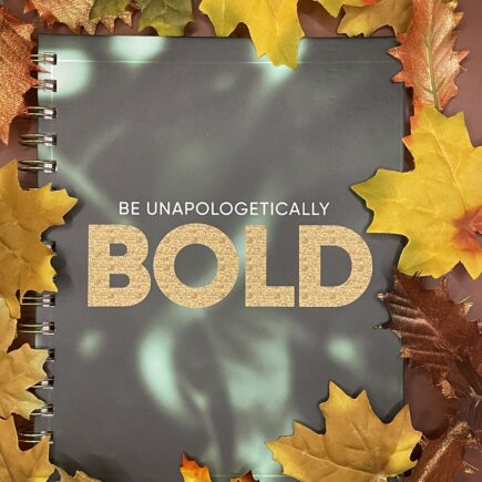 A dark green notebook with light green elements. The text "Be Unapologetically Bold" in gold.