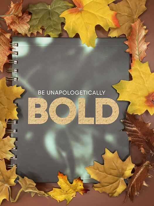 A dark green notebook with light green elements. The text "Be Unapologetically Bold" in gold.