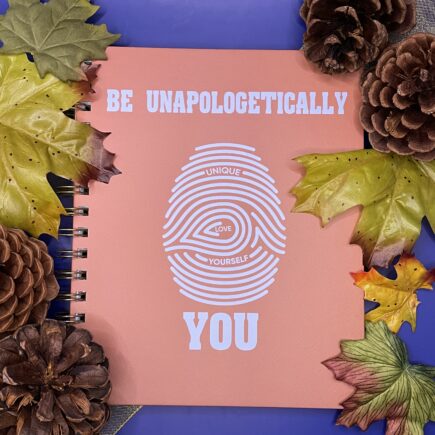 Light orange notebook with light blue text that reads "Be Unapologetically You" and a light blue fingerprint