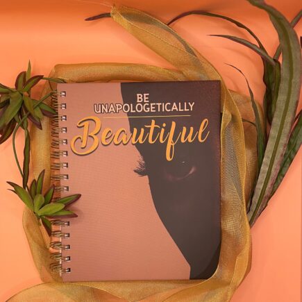 Tan notebook with dark skin black girl's face with the text "Be Unapologetically Beautiful" in dark yellow