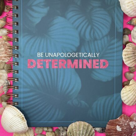 A turquoise blue notebook with light blue shells. The words "Be Unapologetically" are small in white and "Determined" in bold hot pink text.