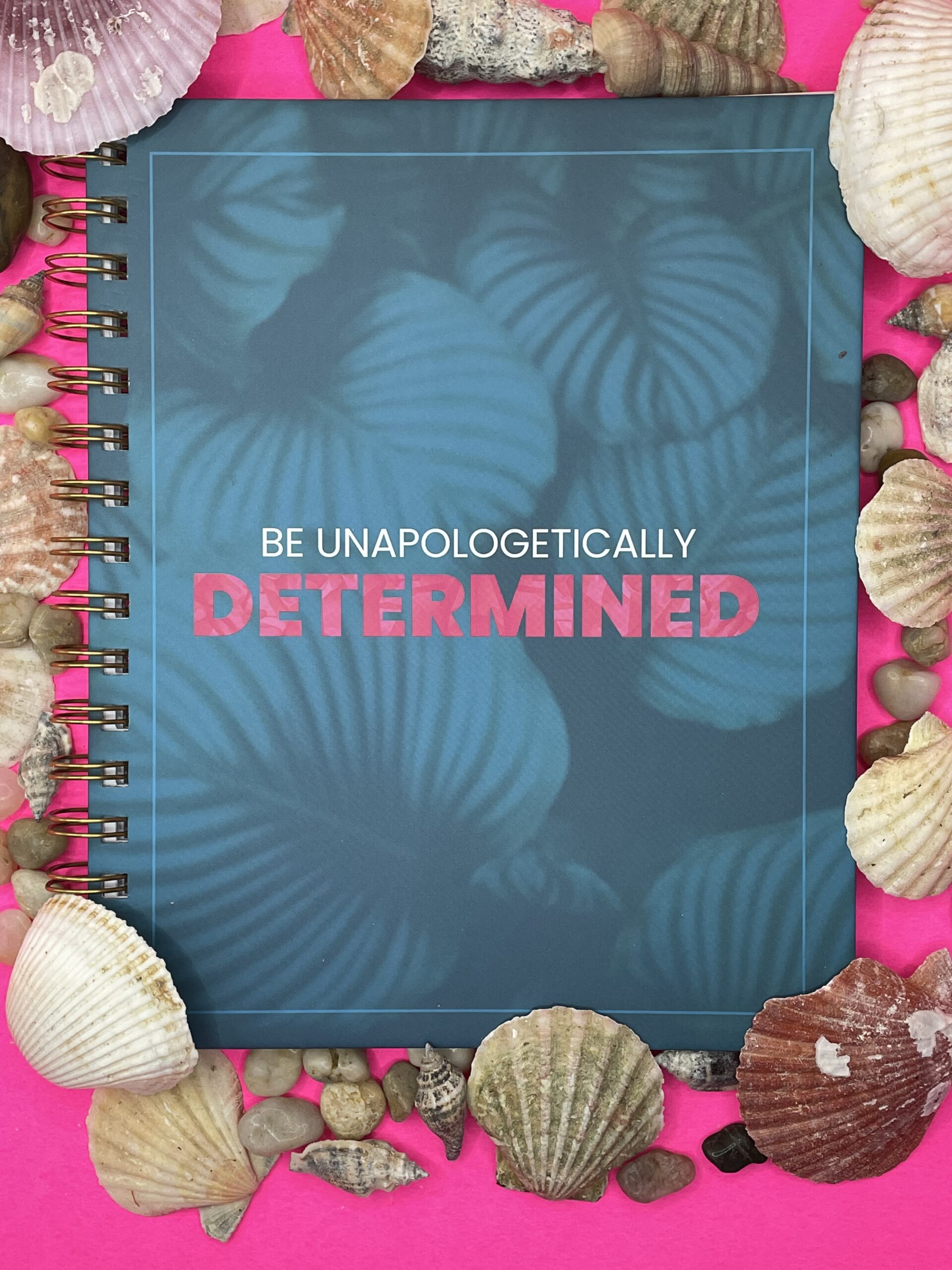 A turquoise blue notebook with light blue shells. The words "Be Unapologetically" are small in white and "Determined" in bold hot pink text.