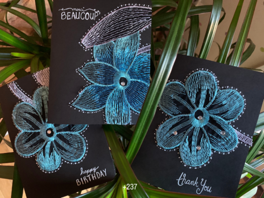 Three black greeting cards with leaves in the background. The cards feature a blue, teal and white embroidered flower, some with decorative gems, with words in white ink like “happy birthday” “thank you” and “merci beaucoup”