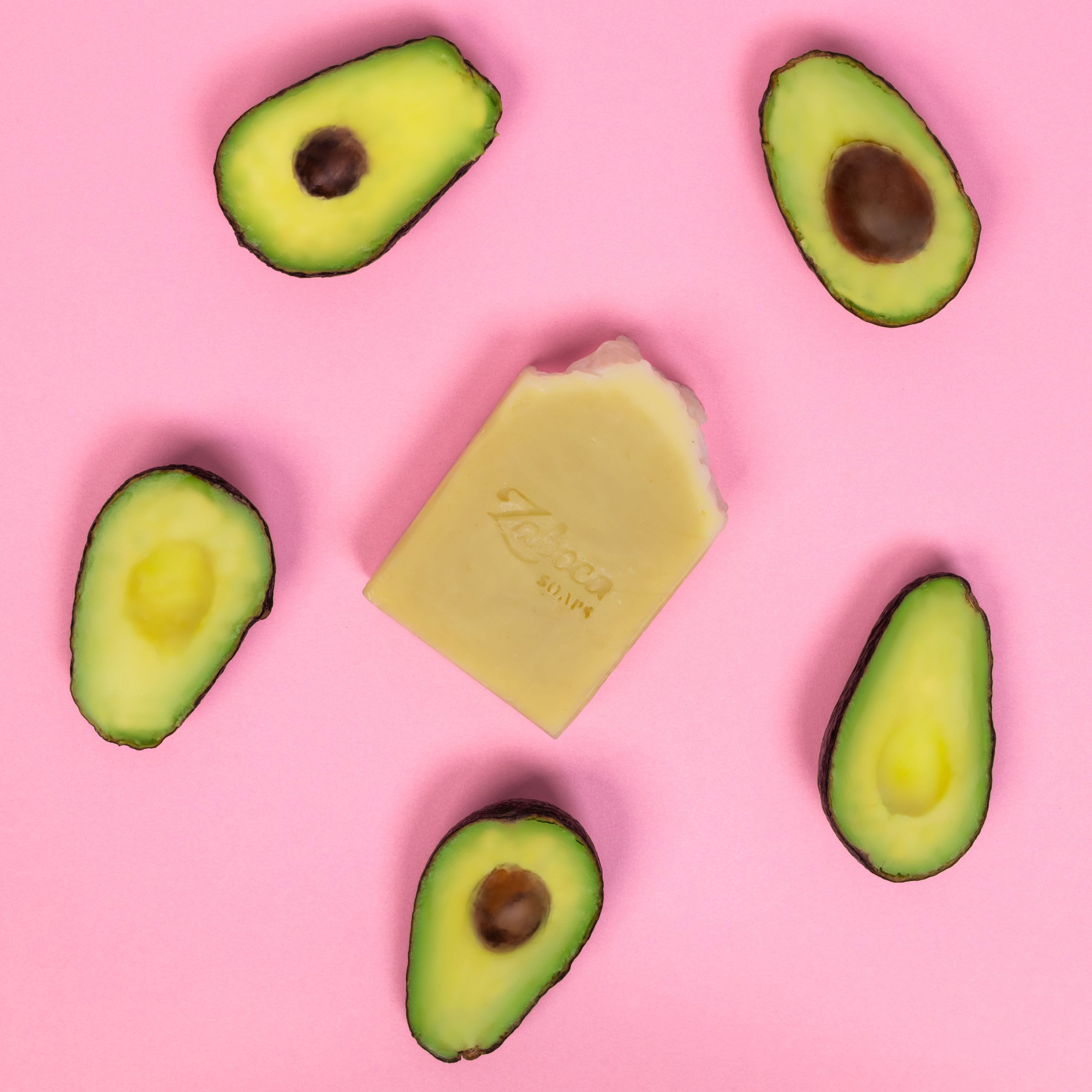 Avocado soap on pink background surrounded by sliced avocados.