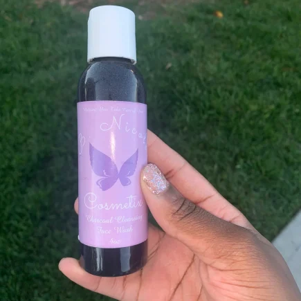 Light purple butterfly logo bottle in room of lush green grass black hand holding the product with pink sparkly nail polish.