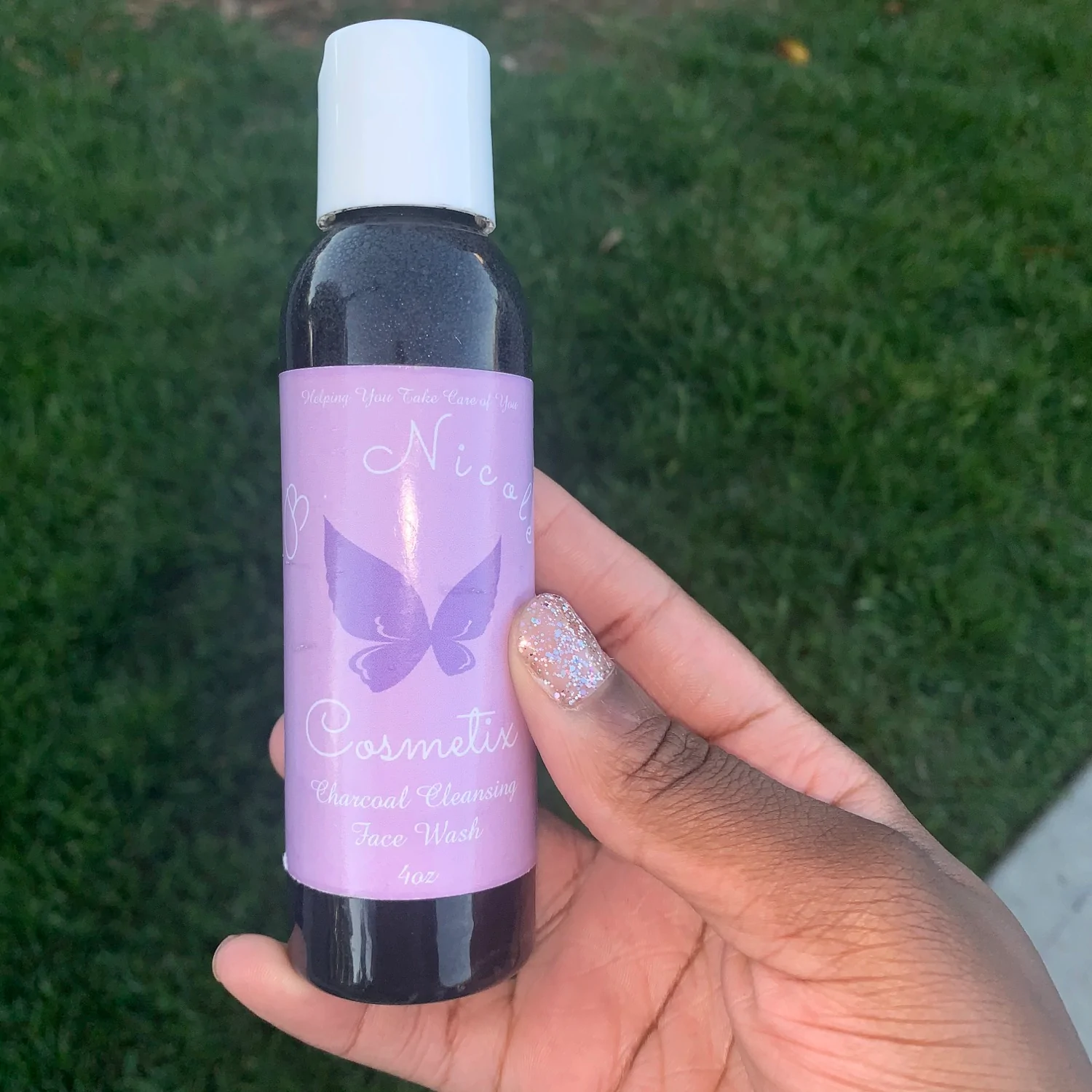 Light purple butterfly logo bottle in room of lush green grass black hand holding the product with pink sparkly nail polish.