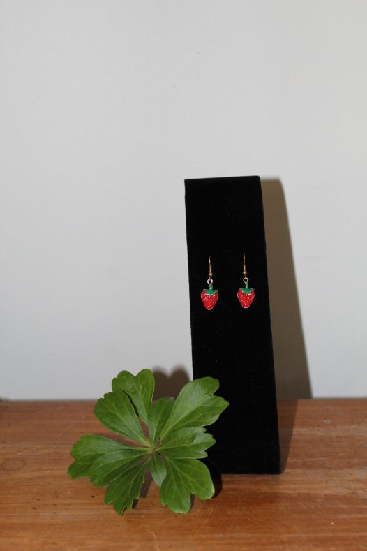 Strawberry dangle earrings on a black stand with a sprig of Japanese pachysandra.