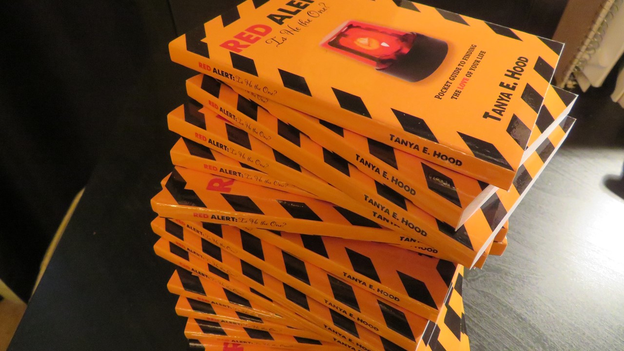 A stack of books with yellow covers and black stripes, featuring the title "RED ALERT" in large red letters, is prominently displayed. The cover design includes a red flashing light, suggesting a possible theme related to warnings or alerts in relationships.