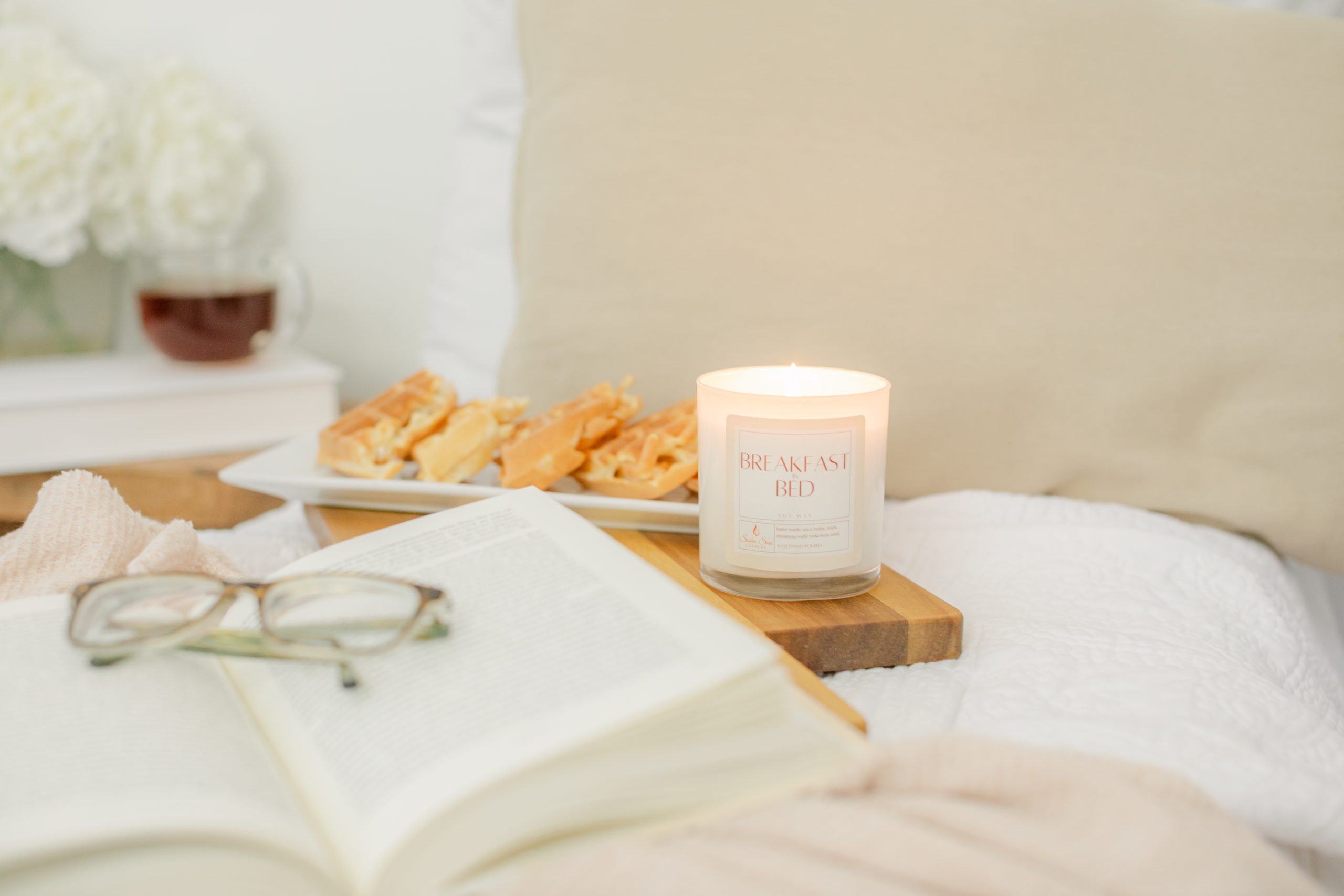 Candle inside white jar next to plate of waffles sitting behind an open book with reading glasses on top.
